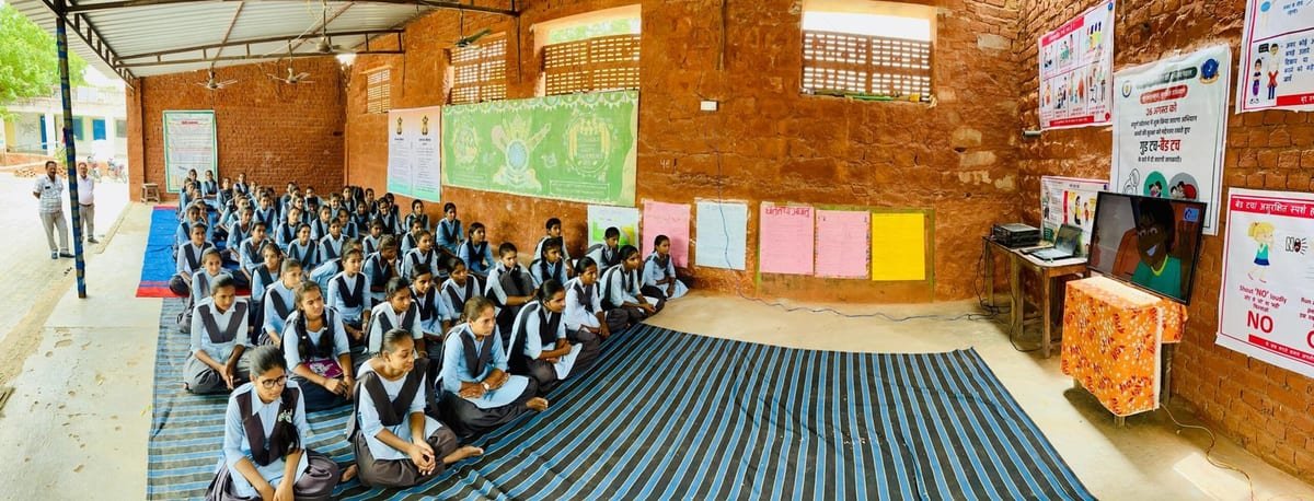 राउमावि जेठन्तरी के बरामदे में दरी पर बैठी छात्राएँ स्मार्ट-टीवी पर दिखाए जा रहे शैक्षिक कार्यक्रम को देखती हुईं; दीवारों पर जागरूकता-पोस्टर एवं शैक्षिक चार्ट